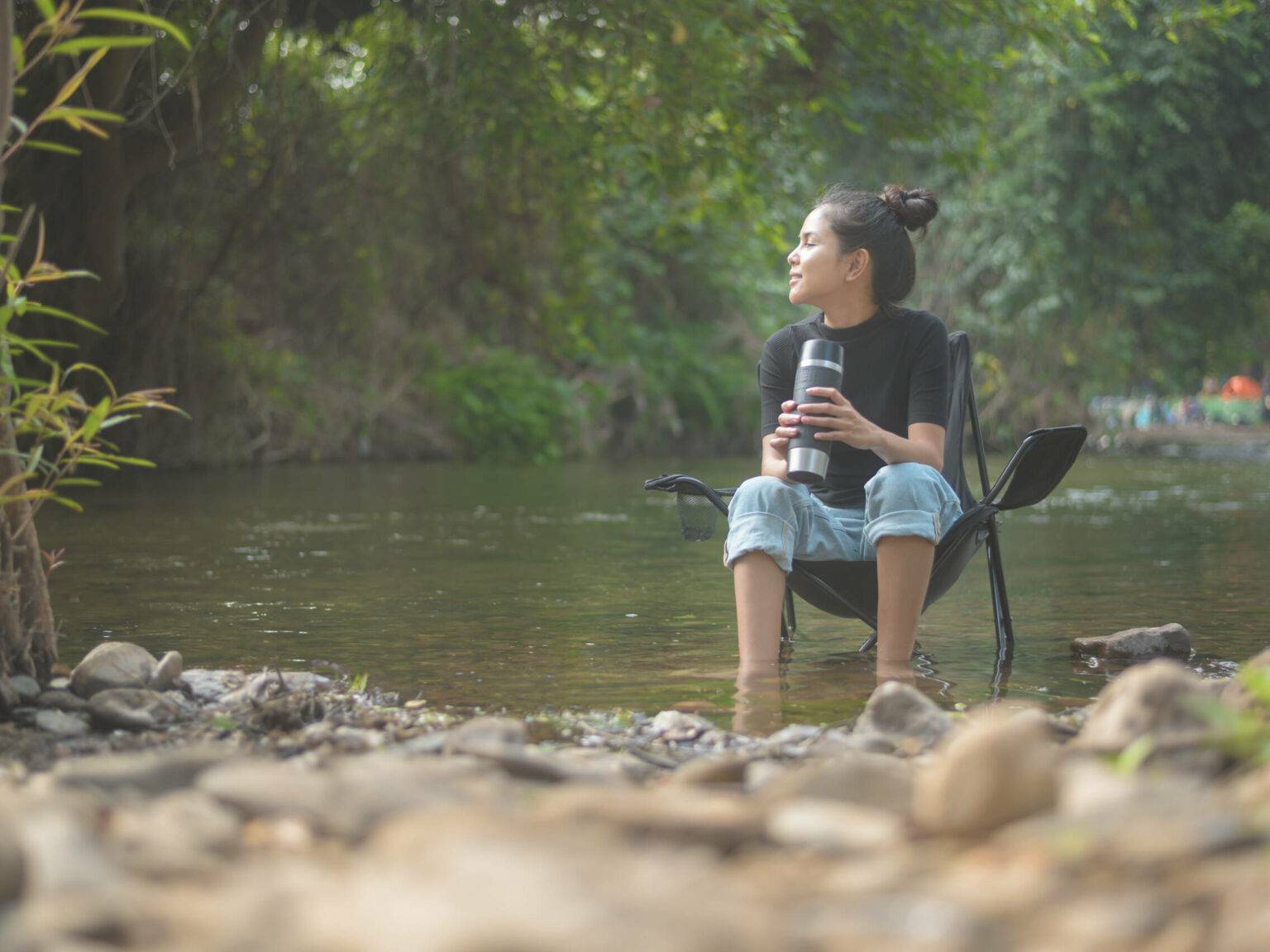 Idyllische Umgebung am See, Frau entspannt im Campingsitz am Wasser mit einem nützlichen Kaffee-Gadget.