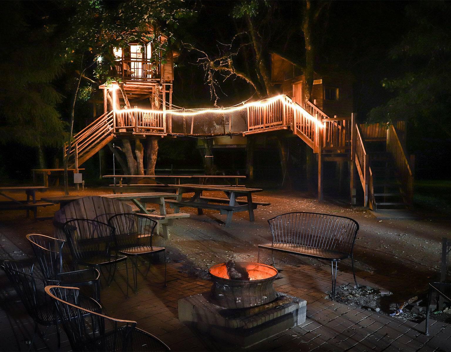 Ein Baumhaus mit Holzsteg und Lichterkette im Hintergrund einer Sitzlounge im freien mit Lagerfeuer.